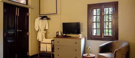 Una habitación de hotel con piso de madera, un televisor sobre una cómoda, una ventana con múltiples divisiones, una silla y un perchero con bata.
