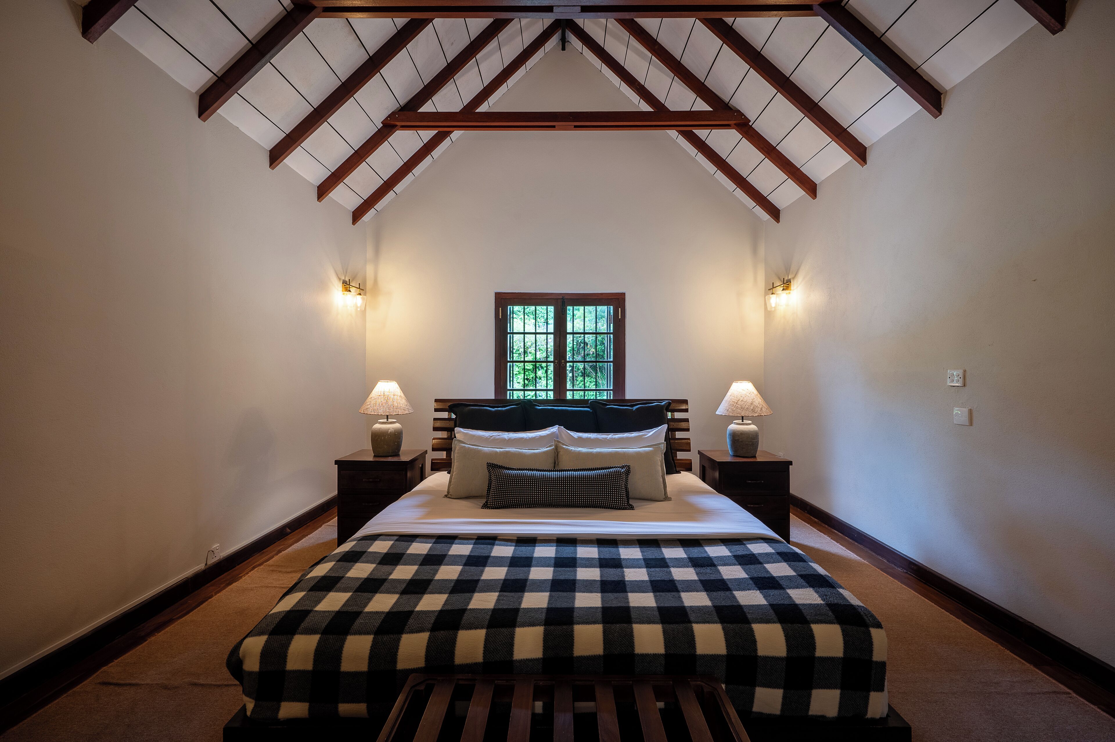 Een slaapkamer met een hoge plafond, een groot bed met een geruit dekbed, twee bedlampen en een raam met uitzicht op groen.