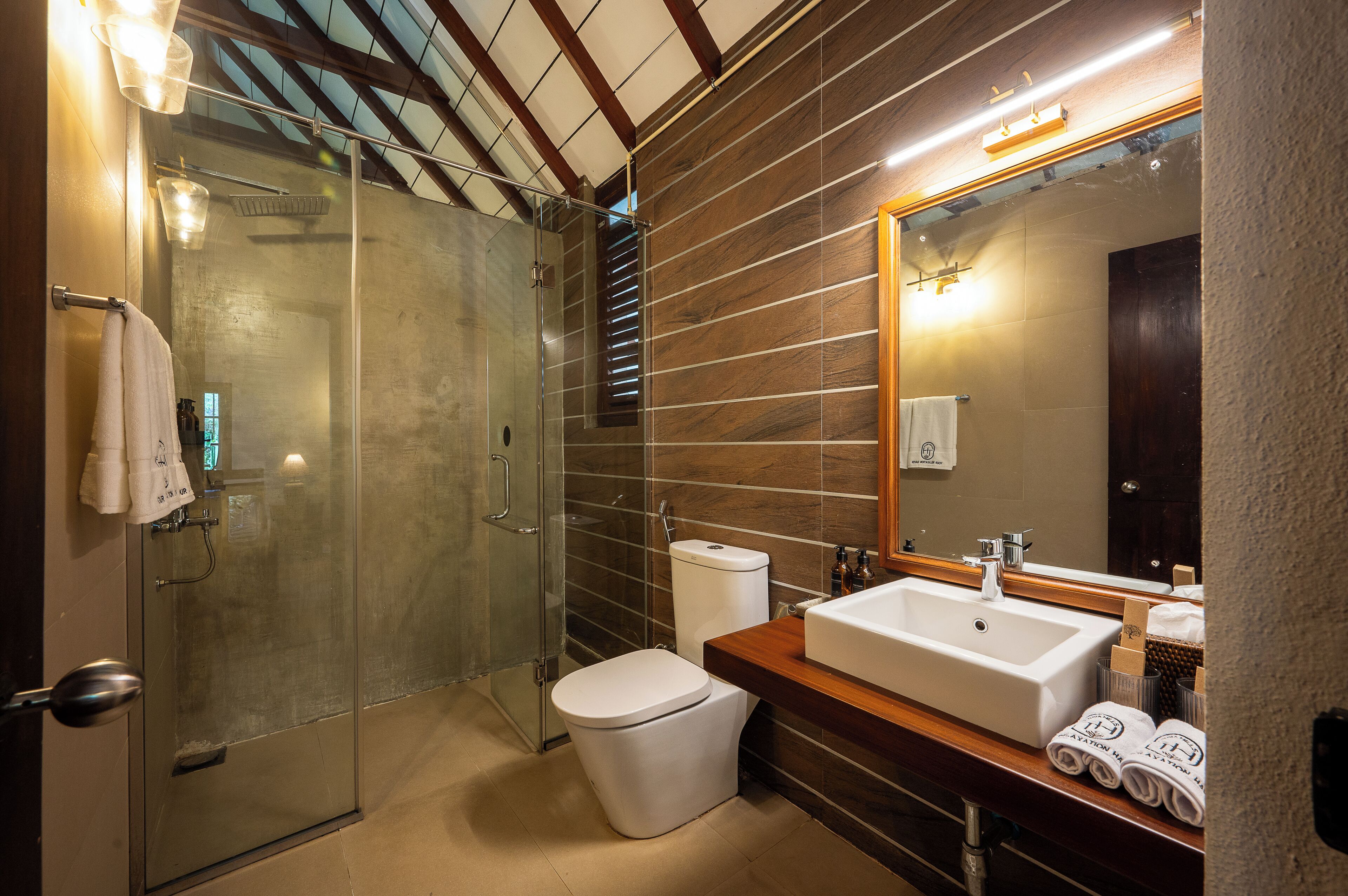Een moderne badkamer met een glazen douchecabine, een wit toilet en een houten wastafelmeubel met een grote spiegel.