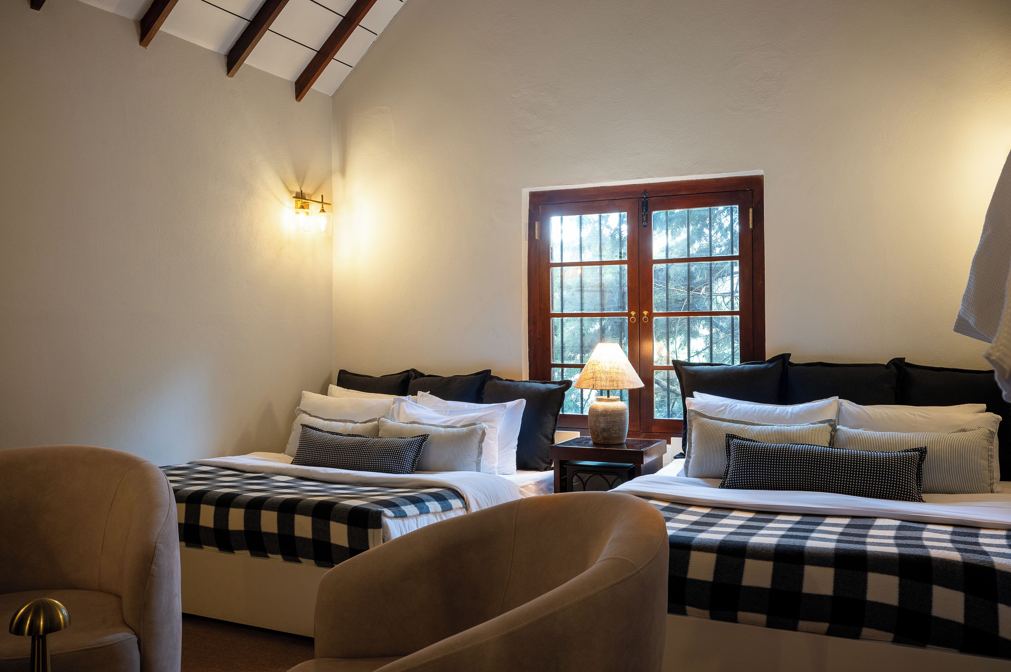 A hotel room with two beds, a chair, a lamp, and a window with a view of trees.