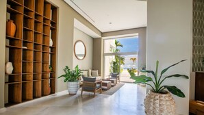 A spacious living area with a large wooden shelving unit, comfortable seating, and large windows offering a view of palm trees and a clear sky. - Beachfront Studio with Balcony, Rooftop Pool & Gym Access (Puerto Vallarta)
