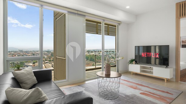 Una sala de estar moderna con un televisor grande, un sofá y vistas de la ciudad a través de amplias ventanas.