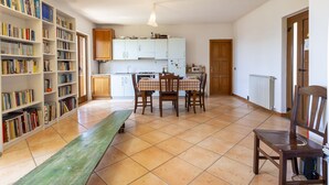 A spacious living area with a dining table, chairs, a bench, a bookshelf, and a kitchen in the background.