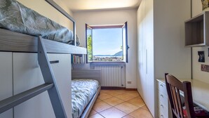 A compact bedroom with a bunk bed, a window with a view of the sea, a radiator, and a small desk.