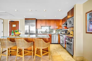 A modern kitchen with wooden cabinets, stainless steel appliances, a marble island, and wicker bar stools. - 2 Honua Kai Villas | Ocean View | Sleeps 14 | Free Car w/ 6+ Nights | ML-1285 (Lahaina)