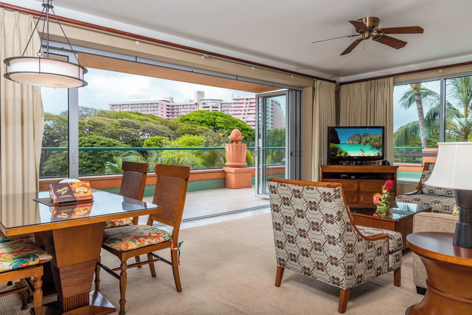 Habitación de hotel con balcón, una mesa de comedor, sillas, sofá, televisor y ventilador de techo.