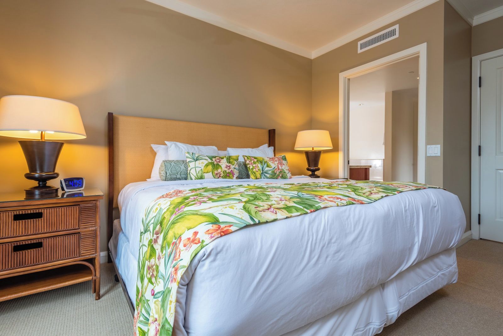 Une chambre d’hôtel comprenant un lit, deux tables de chevet, une petite table, une porte et une bouche d’aération fixée au mur.