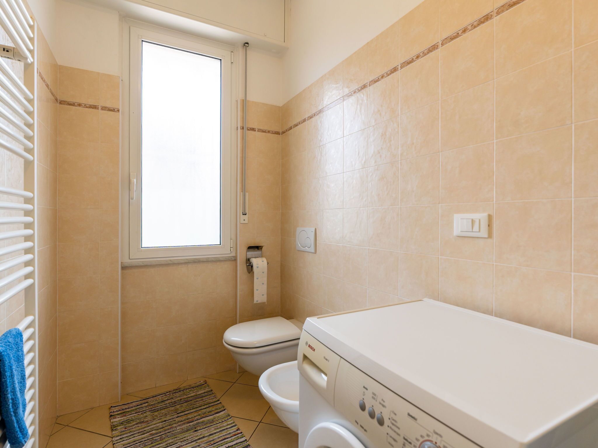Un bagno piccolo e pulito, con piastrelle beige, un water, una lavatrice e una finestra.