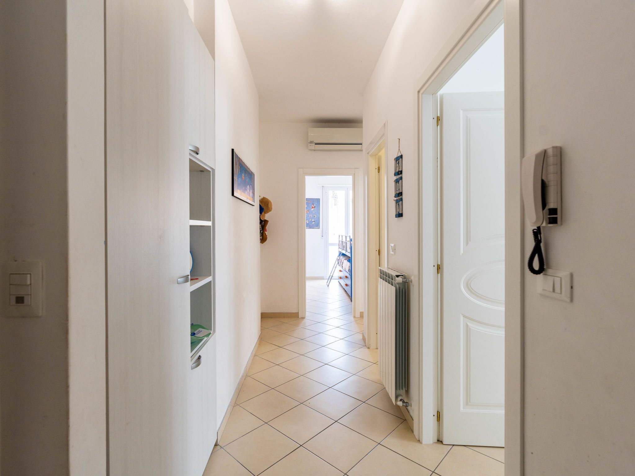 Ein Flur mit weißen Wänden, einem gefliesten Boden und einer Tür rechts. An der Wand befindet sich ein Telefon, links ein Heizkörper.