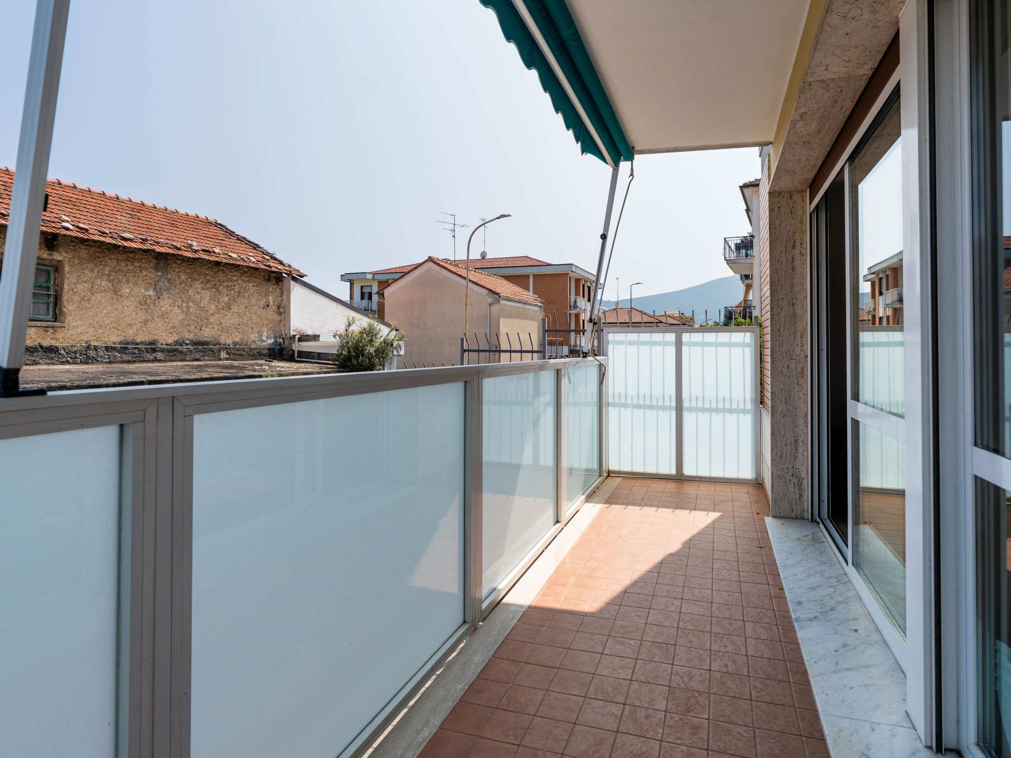Een balkon met betegelde vloer, een glazen deur en uitzicht op omliggende gebouwen.