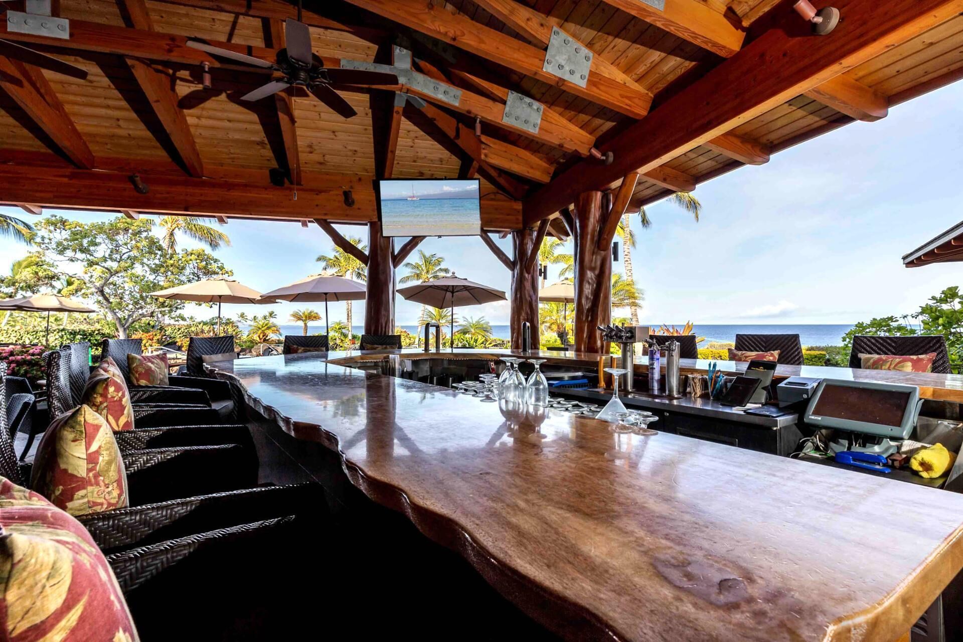 An outdoor bar area with wooden beams, a ceiling fan, and a view of the ocean.