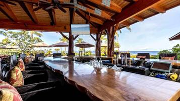 An outdoor bar area with wooden beams, a ceiling fan, and a view of the ocean.