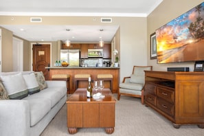 A living room with a sofa, coffee table, TV, and a kitchen area in the background.