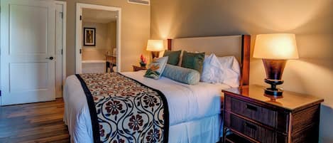 Quarto com cama, abajures, mesa de cabeceira, cabeceira de madeira e porta que dá para outro cômodo.