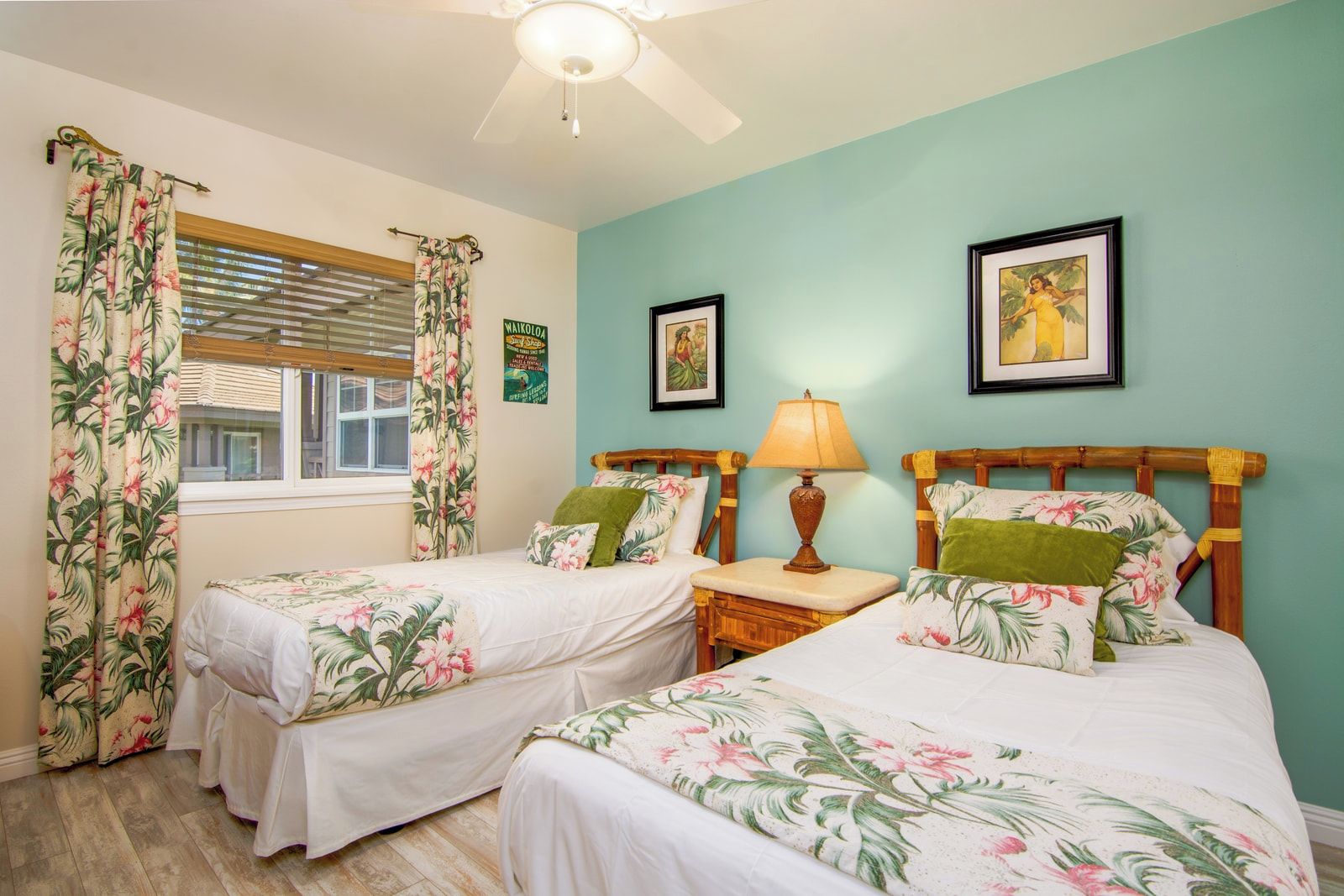 A bedroom with two single beds, a nightstand with a lamp, a window with blinds, and a wall with framed pictures.