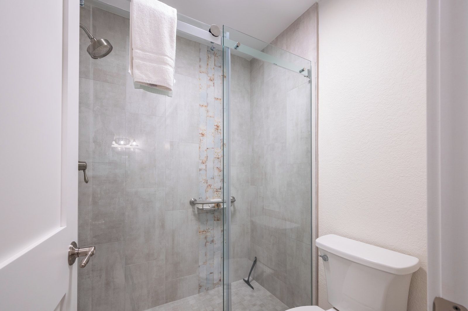 En glasafskærmet brusekabine med et hvidt håndklæde hængende, en glashyde og et hvidt toilet med hvid tank.