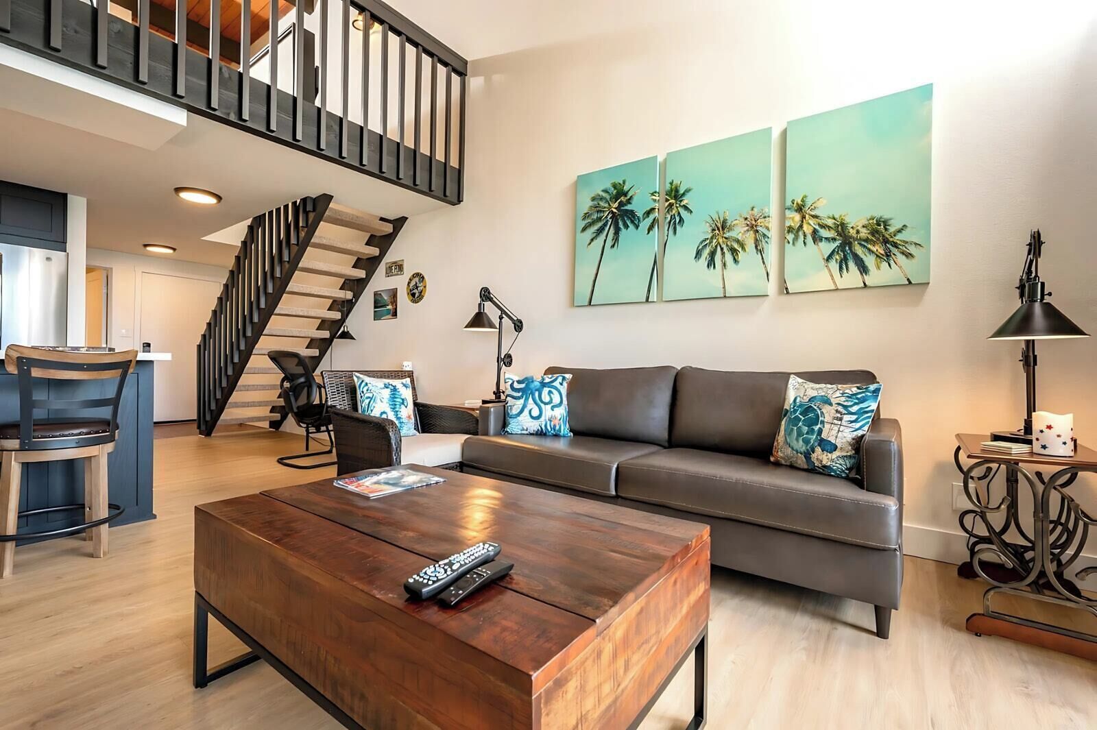 A modern living room with a wooden coffee table, a grey sofa with patterned cushions, a black chair, a staircase, and a wall with three palm tree paintings.