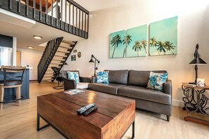 A modern living room with a wooden coffee table, a grey sofa with patterned cushions, a black chair, a staircase, and a wall with three palm tree paintings.