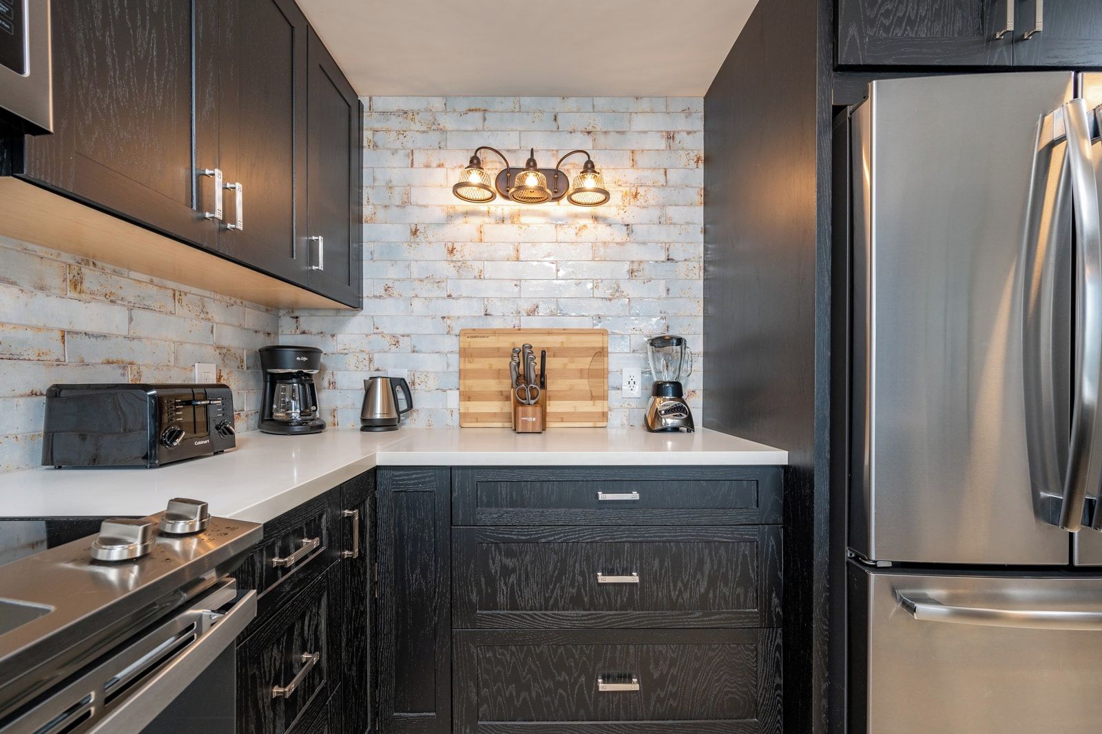 A compact kitchen with dark cabinetry, a stainless steel refrigerator, a coffee maker, a toaster, and a wooden cutting board.