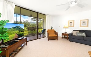 A living room with a TV, sofa, armchair, and a balcony with a view of greenery.