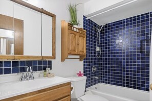 A bathroom with a white sink, wooden cabinets, a mirror, a shower with blue tiles, and a toilet. - Kuilima Estates | Oceanfront 3 BR, Sleeps 10 | Car Incl. w/ 6+ Nights | KUL ML-1303 by KBM (Kahuku)
