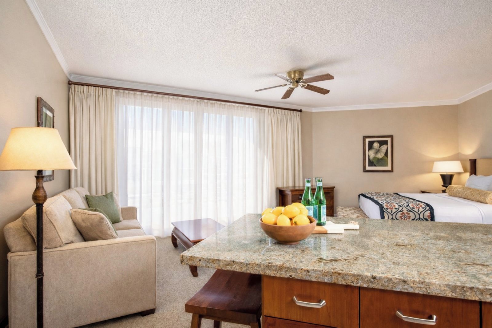 Habitación de hotel con cama, mesa de comedor, zona de cocina con encimera de granito, ventilador de techo y ventana con cortinas.