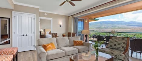 A spacious living area with a sofa, armchair, and coffee table, a ceiling fan, and a balcony with a scenic view.