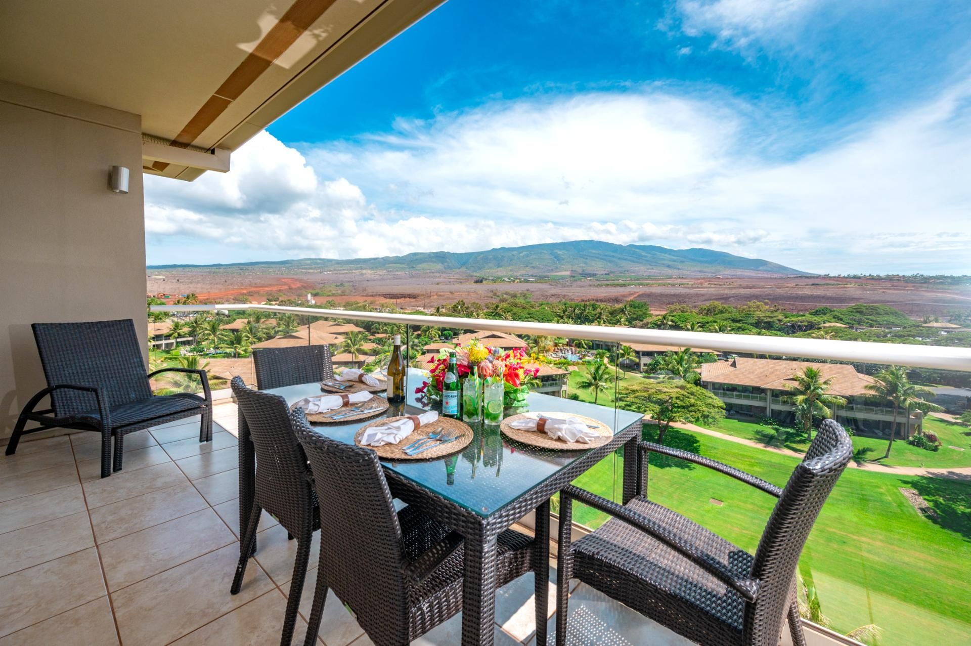 Un balcón con mobiliario de mimbre y una mesa dispuesta para comer, con vistas a un paisaje escénico con montañas y vegetación.