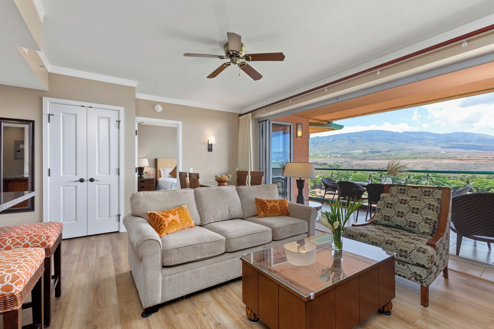 A spacious living area with a sofa, armchair, and coffee table, a ceiling fan, and a balcony with a scenic view.