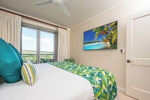 Une chambre d’hôtel avec un lit, un tableau représentant une plage tropicale, un ventilateur de plafond et une vue sur le paysage par la fenêtre.