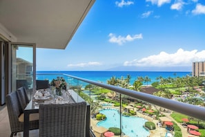 A balcony with a dining table, chairs, and a view of a resort pool and ocean.