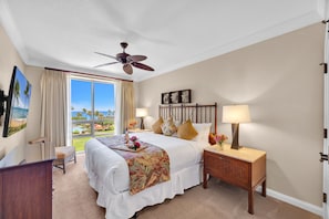 A hotel room with a bed, a desk, a chair, a nightstand, a lamp, a ceiling fan, and a large window with a view of a pool and palm trees. - Honua Kai | Ocean View 5 BR, Sleeps 16 | Car Incl. w/ 6+ Nights | HKH ML-1321 by KBM (Lahaina)