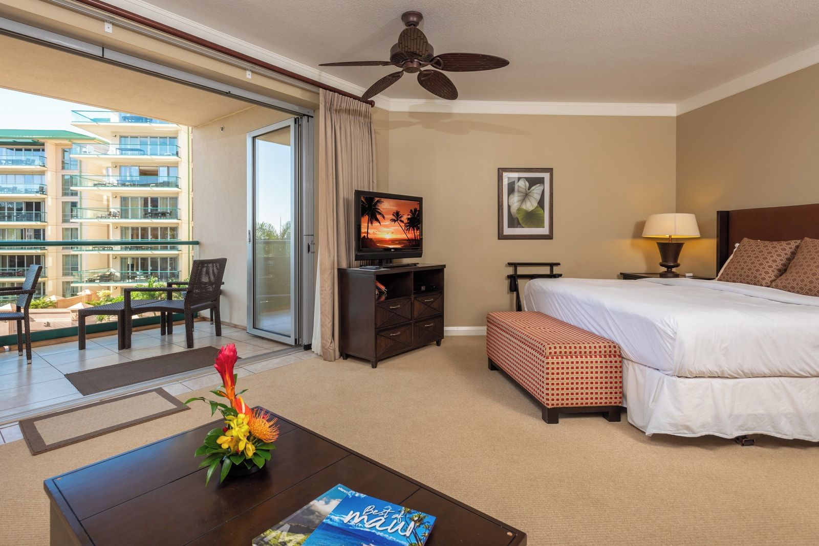 A hotel room with a bed, a television, a desk with a magazine, a coffee table with flowers, and a balcony with chairs and a view of the building.
