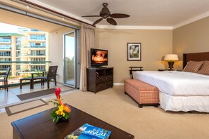 Habitación de hotel con cama, televisor, escritorio con un libro, mesa de centro con flores y balcón con sillas y vista a un edificio.