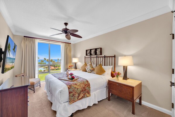 Habitación de hotel con cama, escritorio, silla, mesita de noche, lámpara, ventilador de techo y ventana grande con vista a una piscina y palmeras.