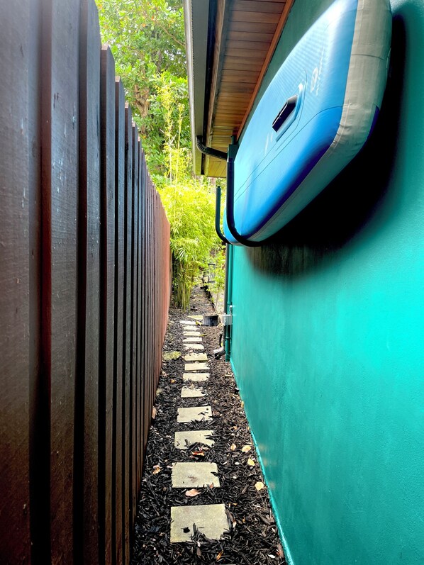 Un étroit sentier bordé d’une clôture en bois d’un côté et d’un mur végétal de l’autre, qui mène à un espace abrité surmonté d’un toit bleu.