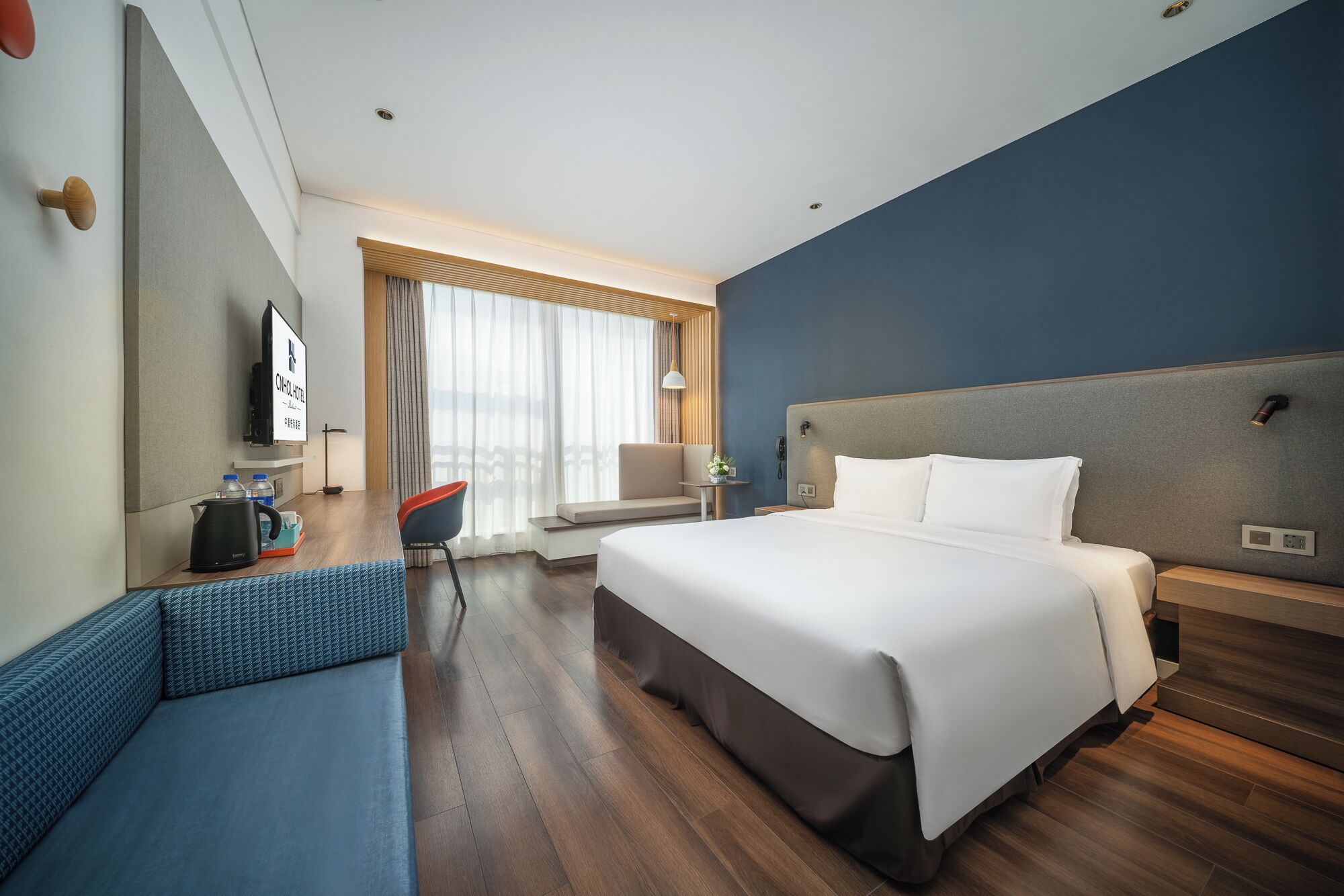 A hotel room with a large bed, a desk with a kettle, a chair, a sofa, a TV, and a window with curtains.