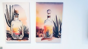 Two wall-mounted paintings of a bottle with a desert landscape, a white chair, and a teal cabinet. - ‘Señorita Sunset’ Guest Fave! Chic Studio Montrose Walkable area! (Houston)