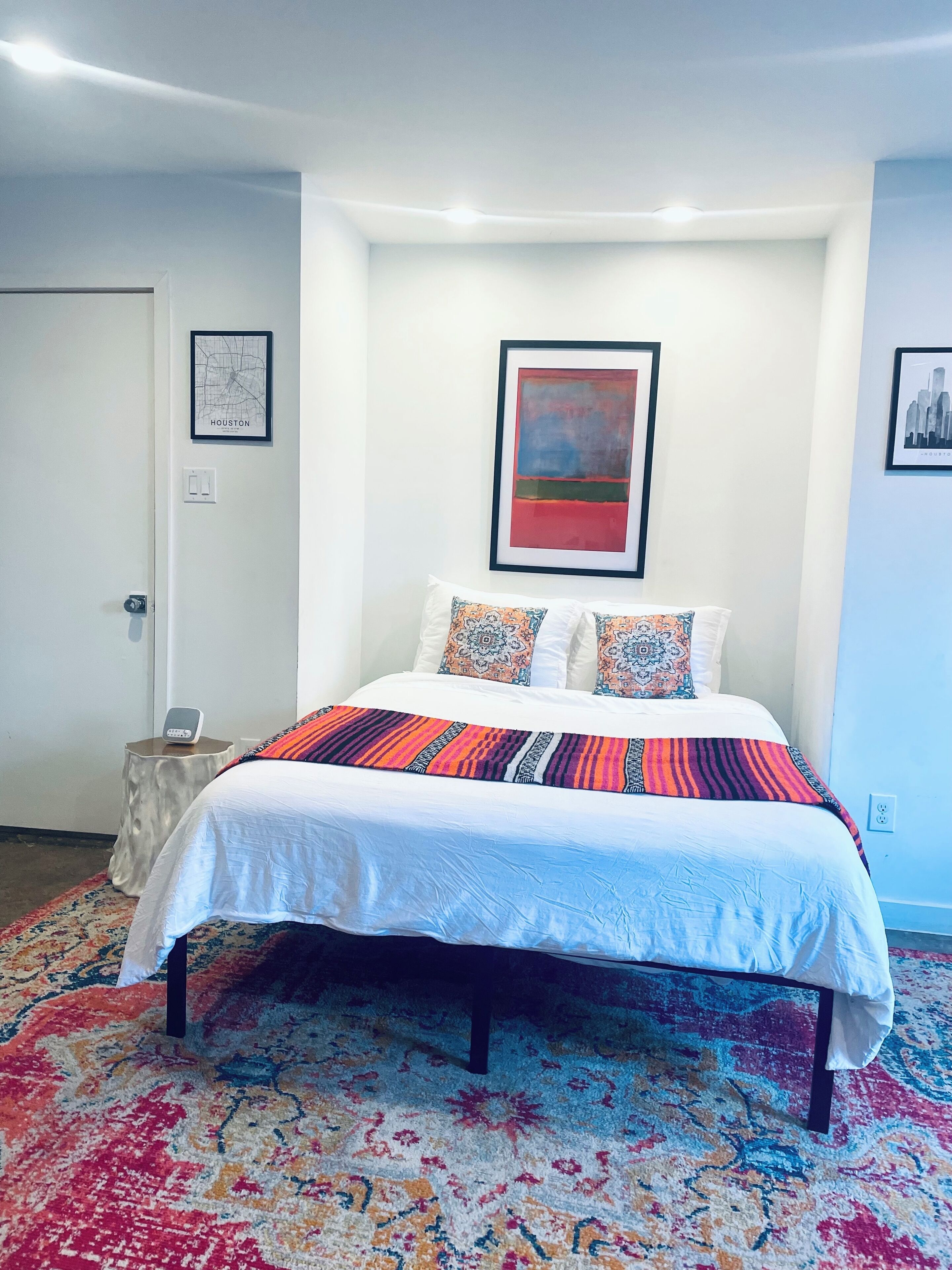 A neatly made bed with a patterned bedspread, a door with a framed map, and a framed abstract painting on the wall.