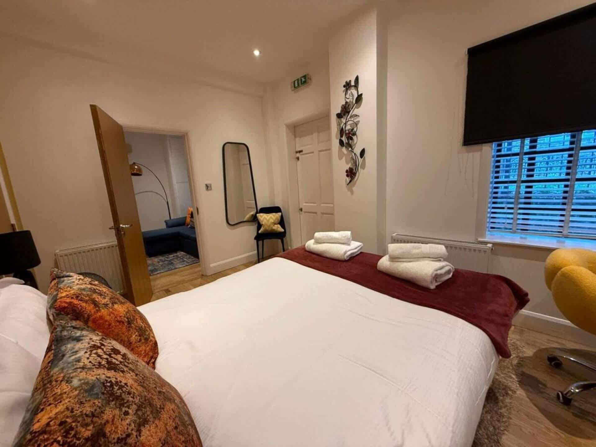 Et hotelværelse med en stor seng, en lænestol, et spejl og et vindue med persienner.
