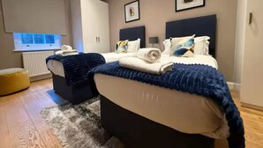 A modern bedroom with two beds, a white wardrobe, a television, and a small yellow ottoman.