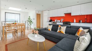 A modern living room with a grey sofa, a dining area with wooden chairs, and a kitchen with white cabinets and a red backsplash. - Bright Angel 2 BR Duplex with Balcony Sleeps 5 (London)