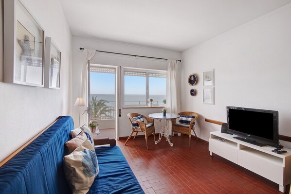 Una habitación con un sofá azul, un televisor sobre un mueble blanco, sillas de mimbre y vistas al mar a través de amplios ventanales.