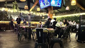 An outdoor dining area with wooden tables and chairs, hanging lights, and a TV screen displaying a show.