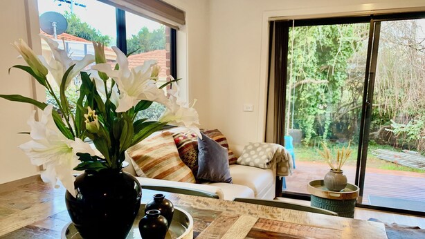 A modern living room with a wooden coffee table, a black vase with white flowers, and a sofa with patterned cushions.
