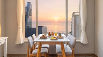 Uma área de refeições com uma mesa posta para o café da manhã, com vista para o pôr do sol sobre o oceano e um edifício moderno.
