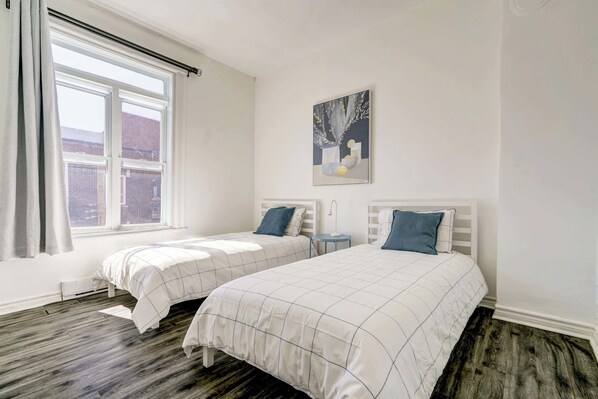 Un dormitorio con dos camas, una ventana con cortinas, una mesita de noche pequeña y un cuadro en la pared.