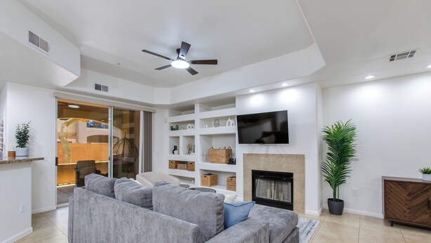 A modern living room with a grey sofa, a flat-screen TV mounted above a fireplace, and a ceiling fan. - Luxury 2-bedroom condo in Scottsdale! (Scottsdale)