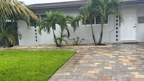 Un camino pavimentado conduce a una casa blanca con techo gris, rodeada por un césped cuidado y palmeras.