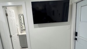 Un pasillo con una puerta blanca, un baño con espejo y perchero, y un televisor de pantalla plana montado en la pared.